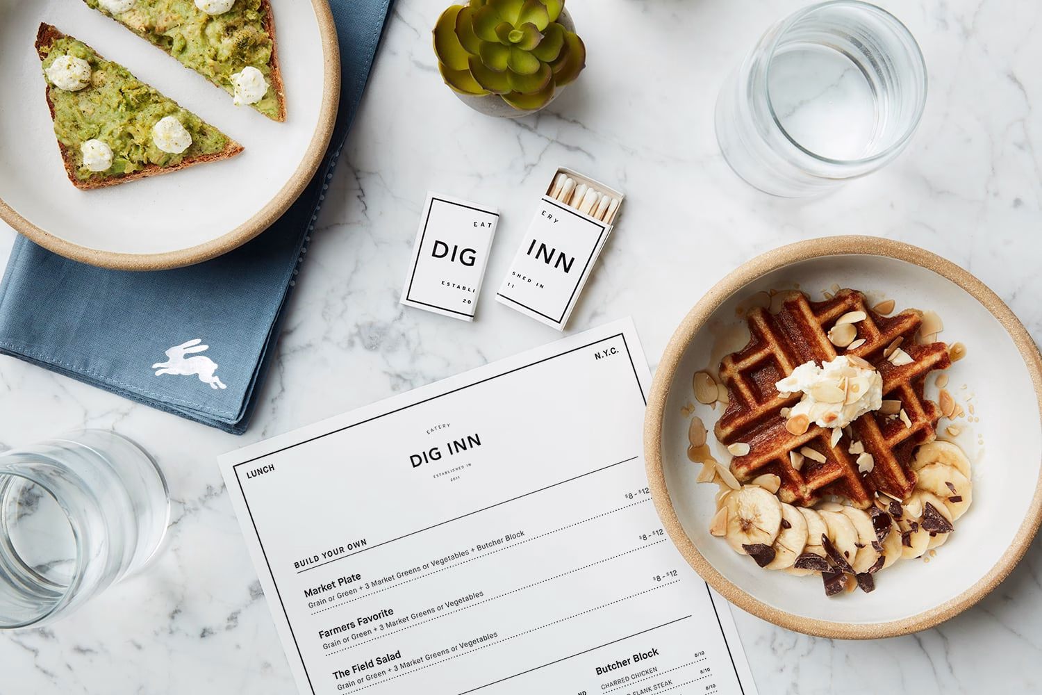 A marble table features a menu, a bowl of waffles with banana, and avocado toast, alongside a small succulent plant.