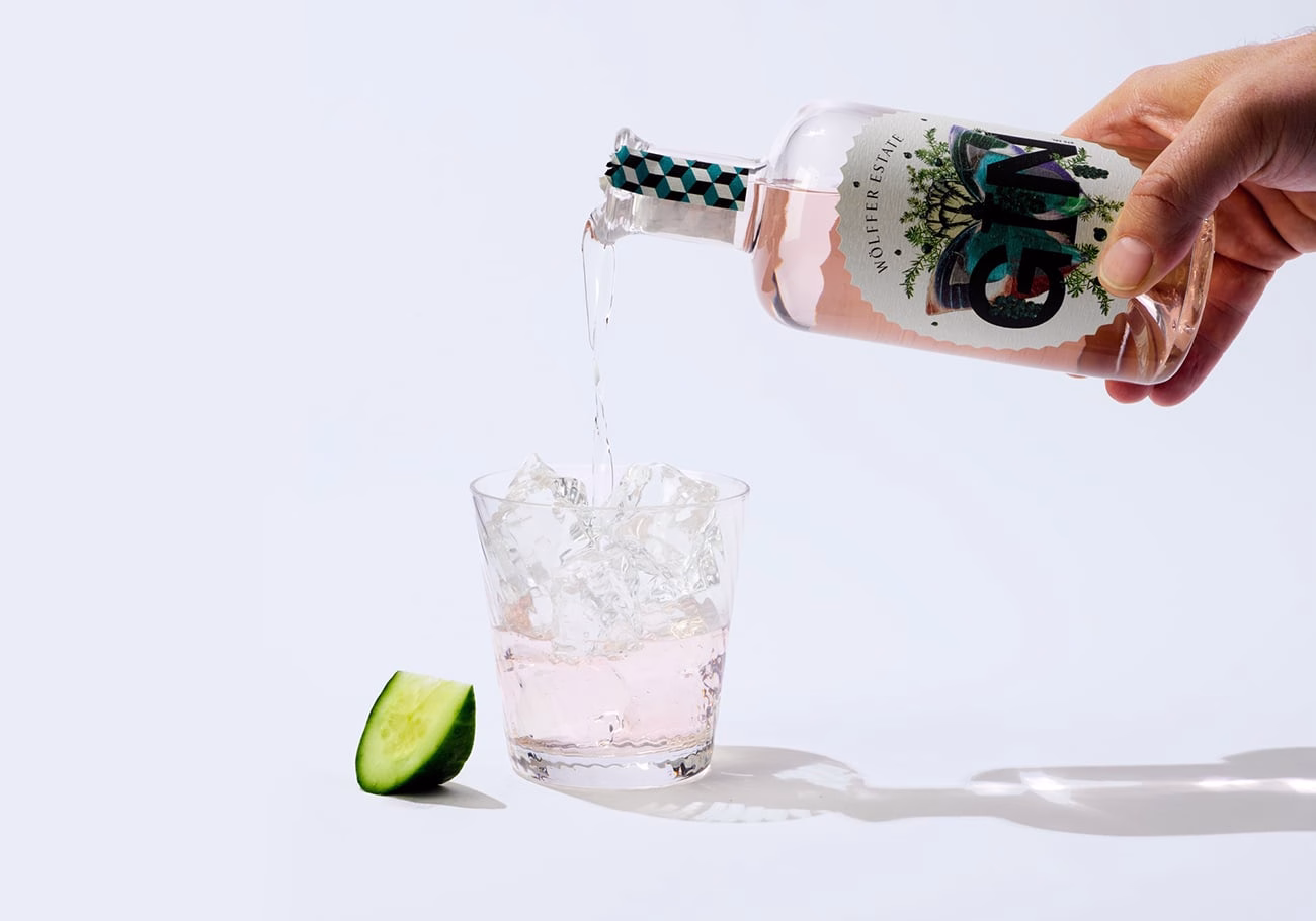 A hand pours gin over ice in a clear glass, with a fresh cucumber slice nearby on a light background.