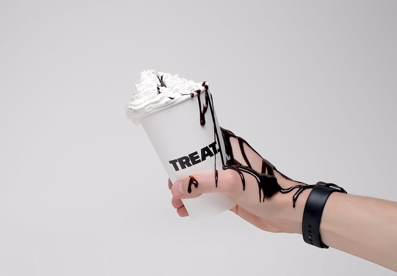 A hand holds a paper cup overflowing with whipped cream and drizzled chocolate sauce, with text reading "TREAT."