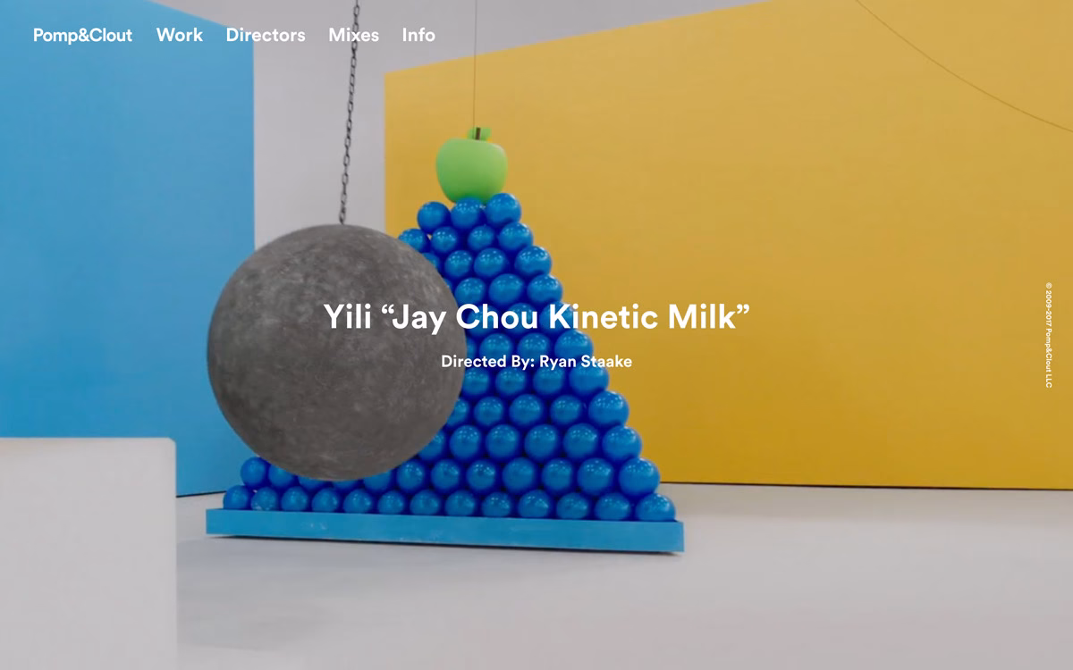A large black sphere hangs in front of a blue and yellow backdrop, resting near a pyramid of blue spheres.