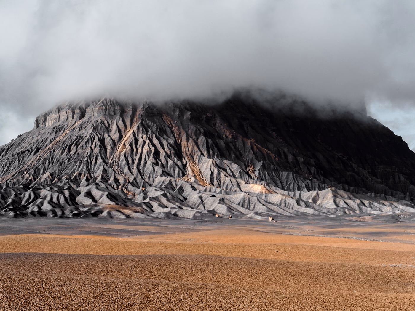 A rugged mountain with sharp ridges rises beneath a cloudy sky, casting shadows over a barren, sandy landscape.