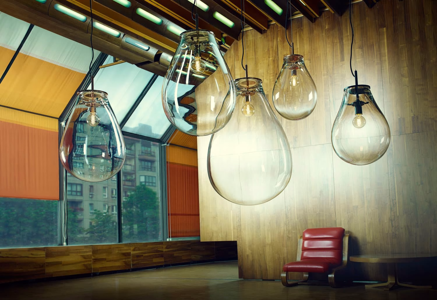 Five glass light fixtures hang above a wooden floor, with a red chair and circular table in a sunlit, modern space.