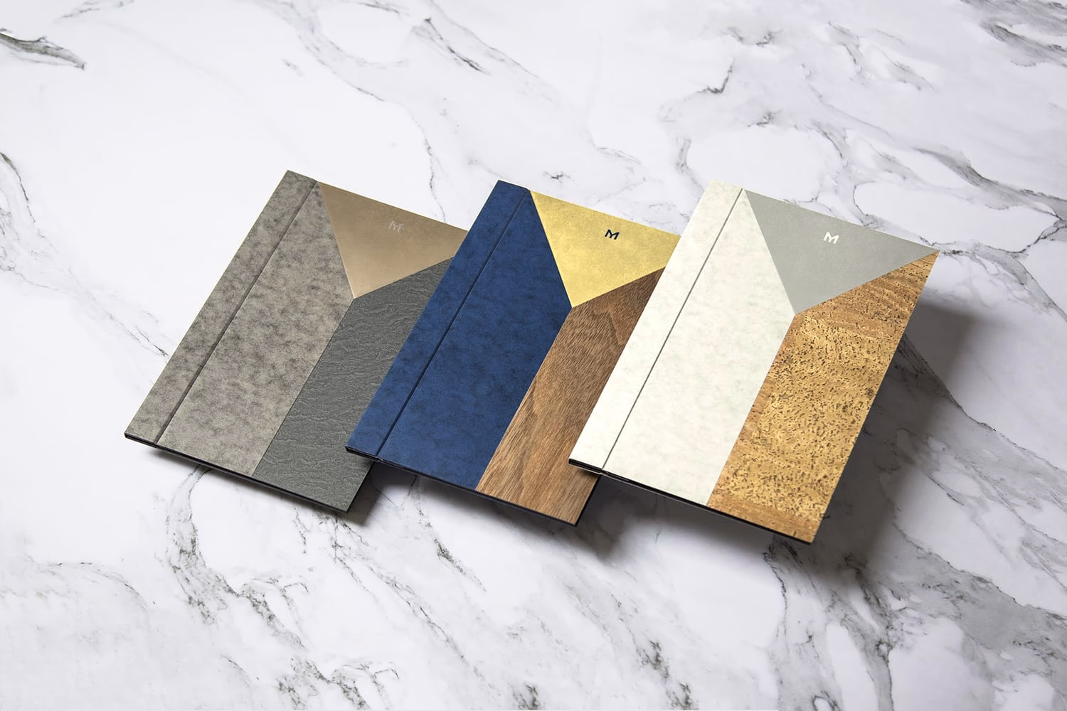 Three notebooks with geometric designs in gray, blue, and light colors displayed on a marble surface.