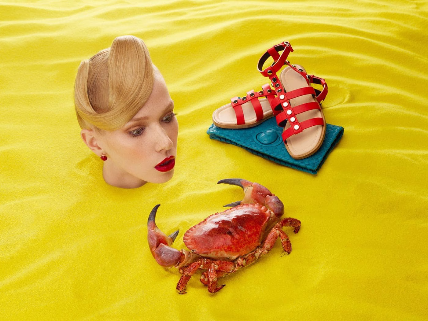 A woman's head emerges from vibrant yellow fabric, next to a red crab and a pair of red-strapped sandals.