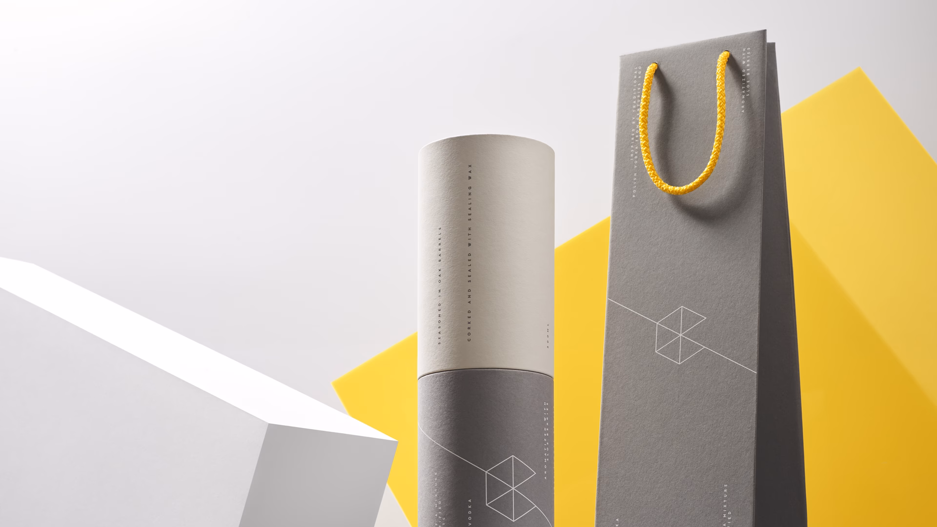 A cylindrical gray and white packaging sits beside a tall gray bag with a yellow rope handle against geometric backdrops.
