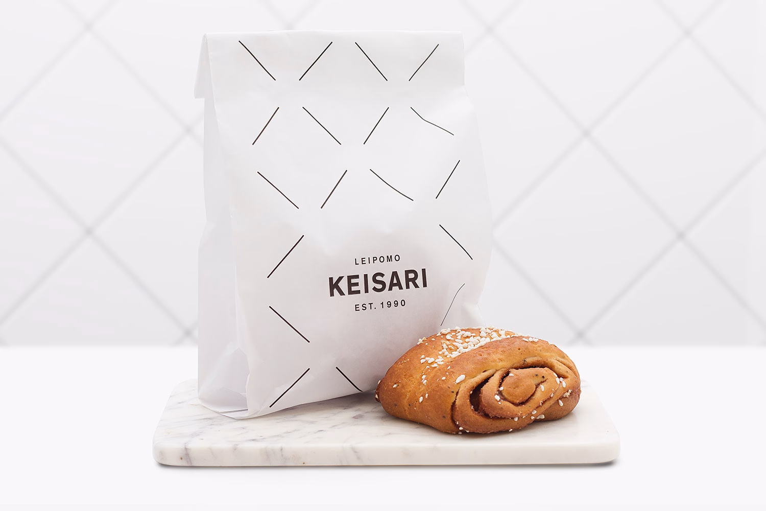 A white bag with black geometric patterns sits on a marble surface next to a cinnamon roll topped with sesame seeds.