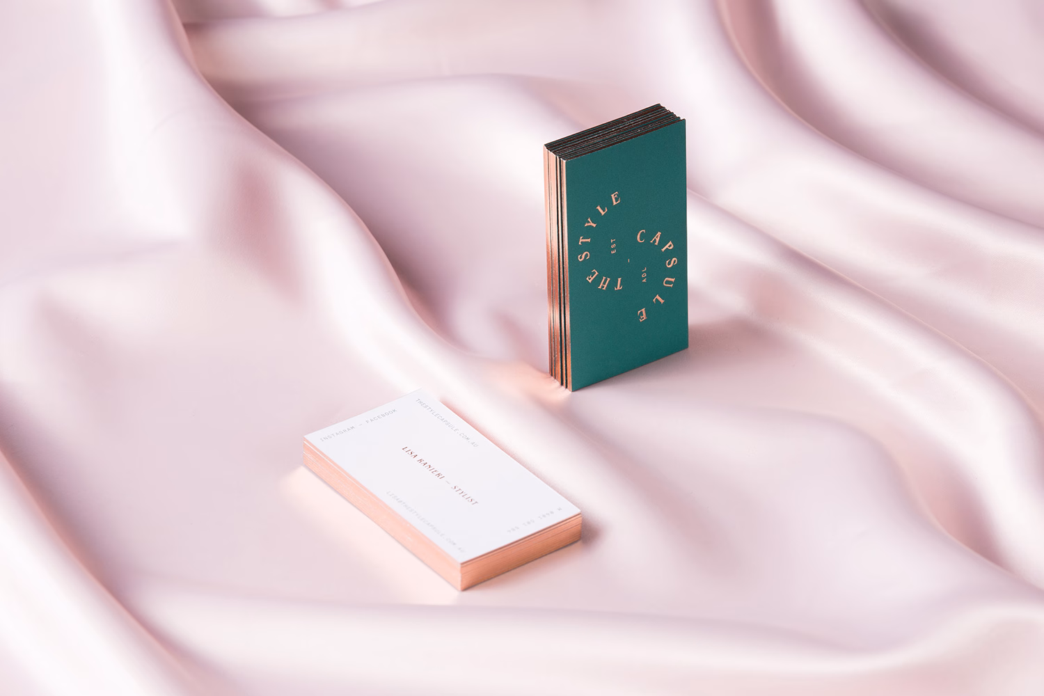 Two business cards resting on soft, pale pink fabric; one features green and gold typography, the other white with a rose gold edge.