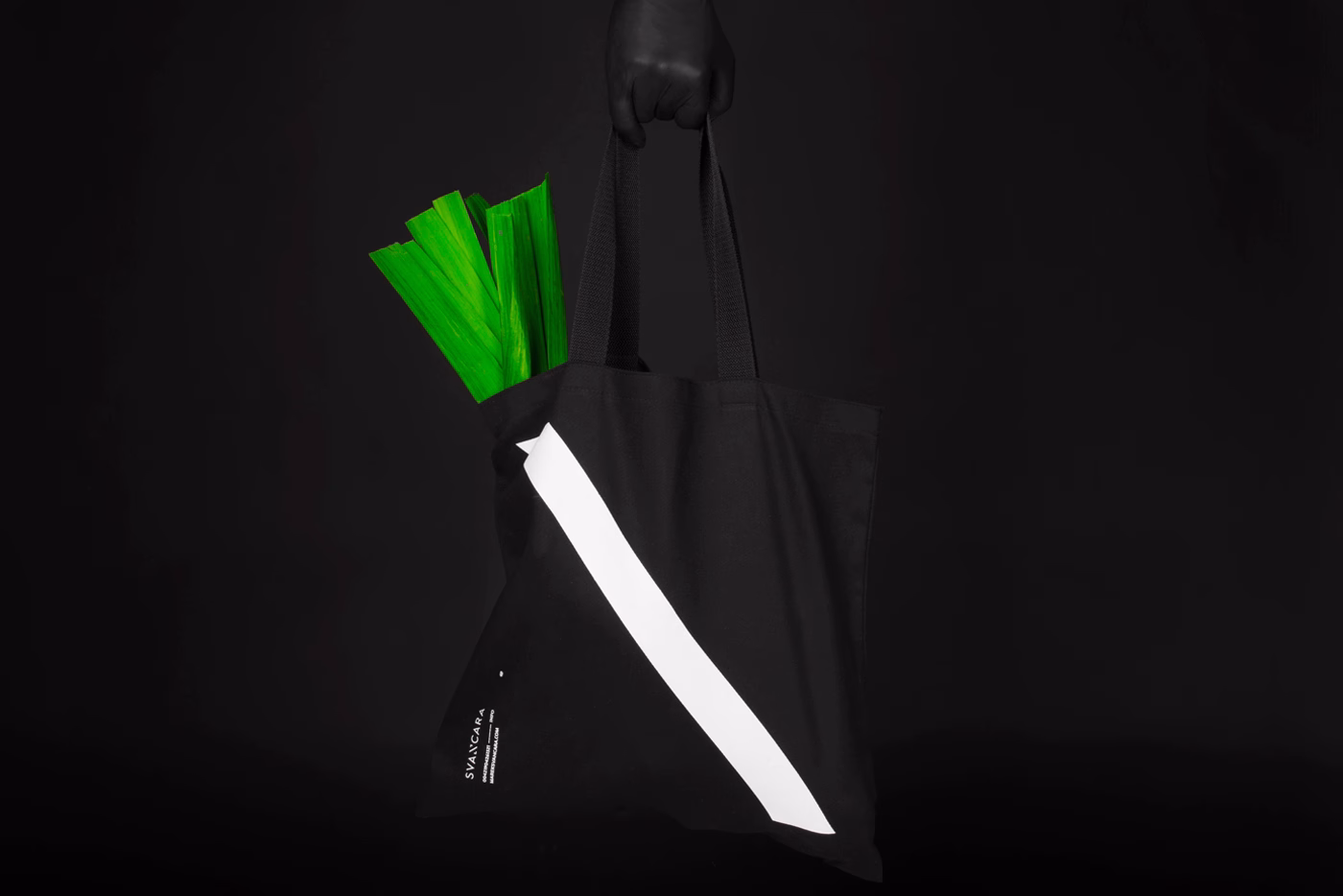 A black tote bag with a white diagonal stripe holds vibrant green stalks, set against a dark background.