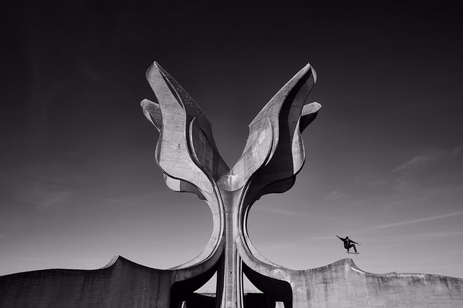 A skateboarder performs a trick mid-air near a large, abstract concrete sculpture against a black and white sky.