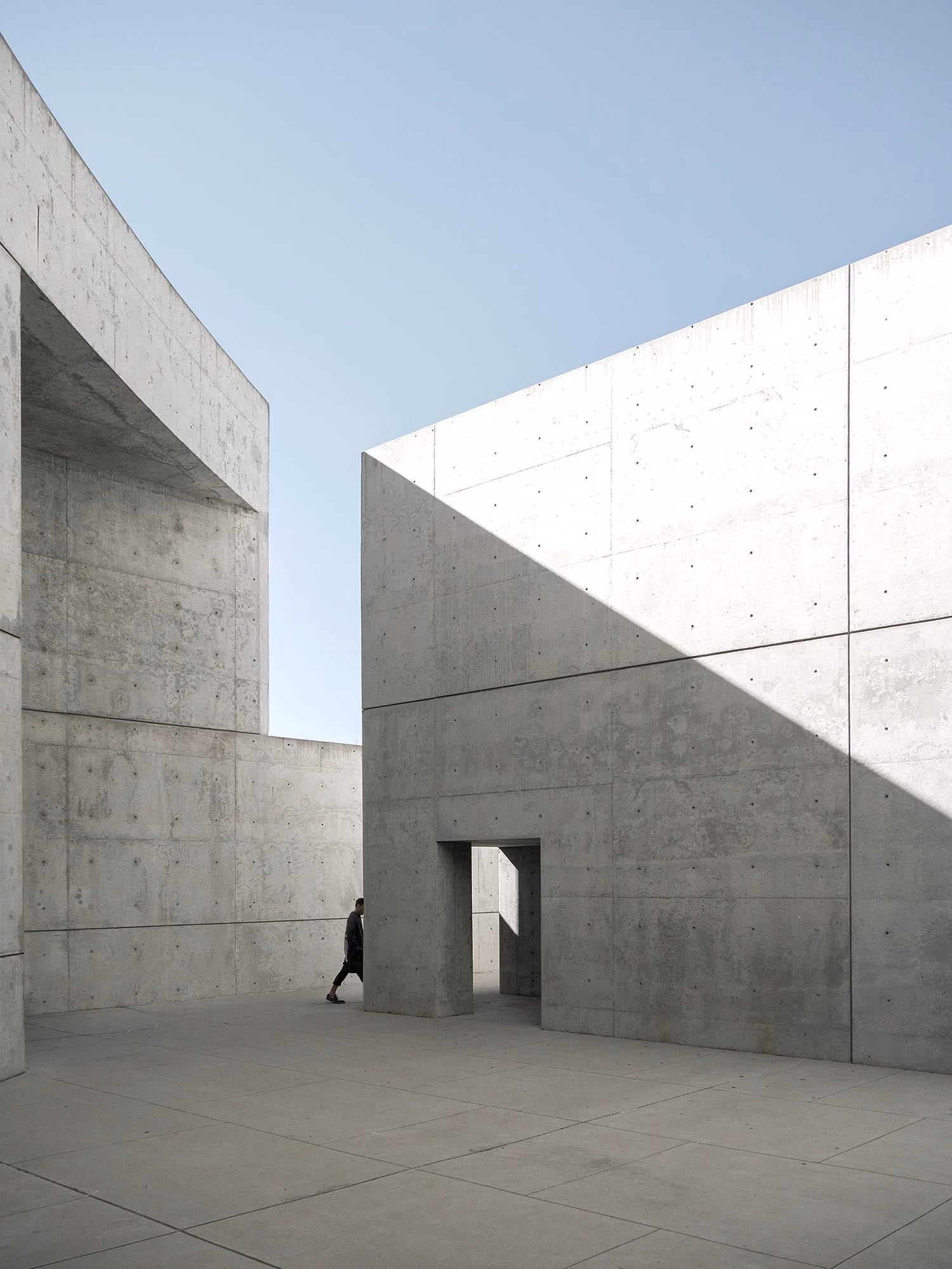 A person walks between angular concrete walls under a clear blue sky, highlighting the structure's sharp geometries.