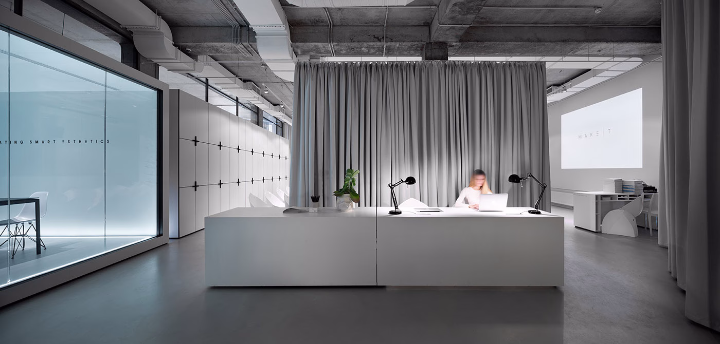 A person works at a sleek white desk in a modern office with soft gray curtains and a minimalist aesthetic.