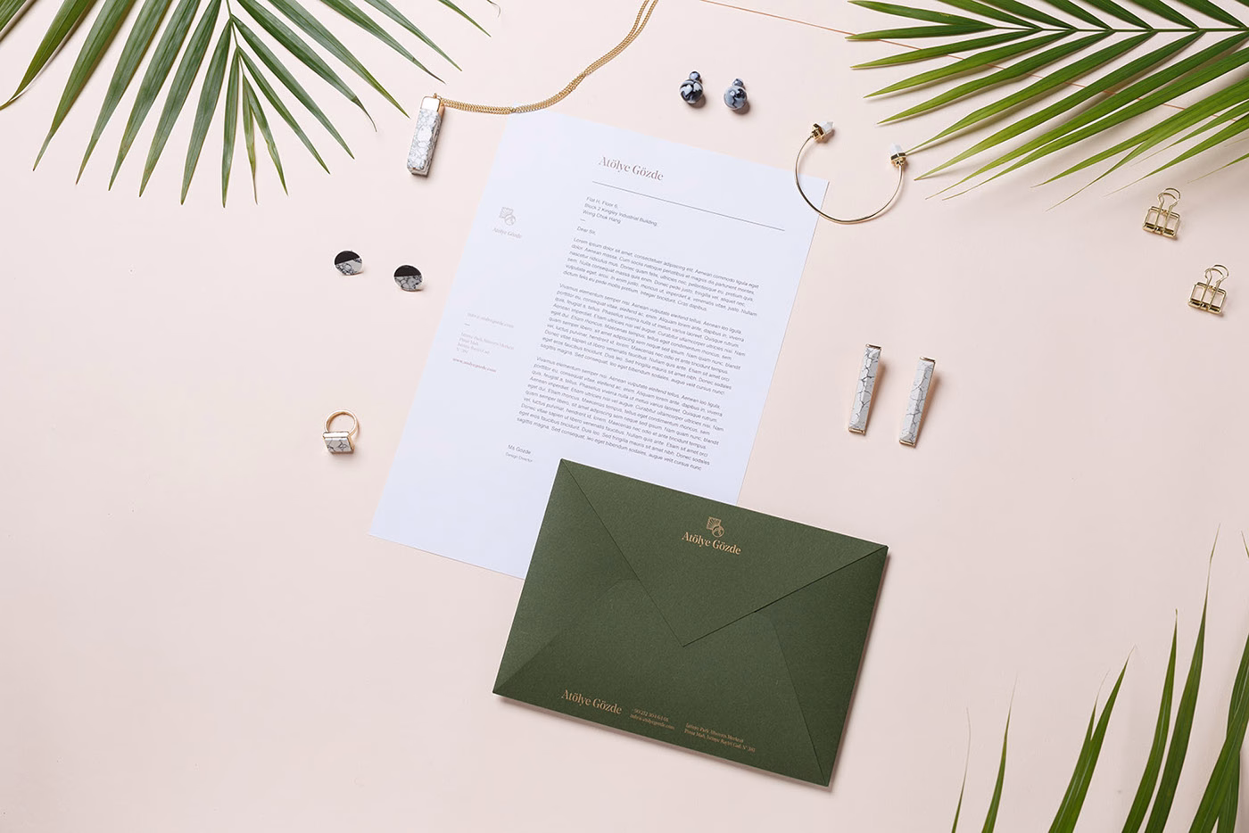 A flat lay features delicate jewelry, a green envelope, and a letter, set against a soft pink background with a palm frond.
