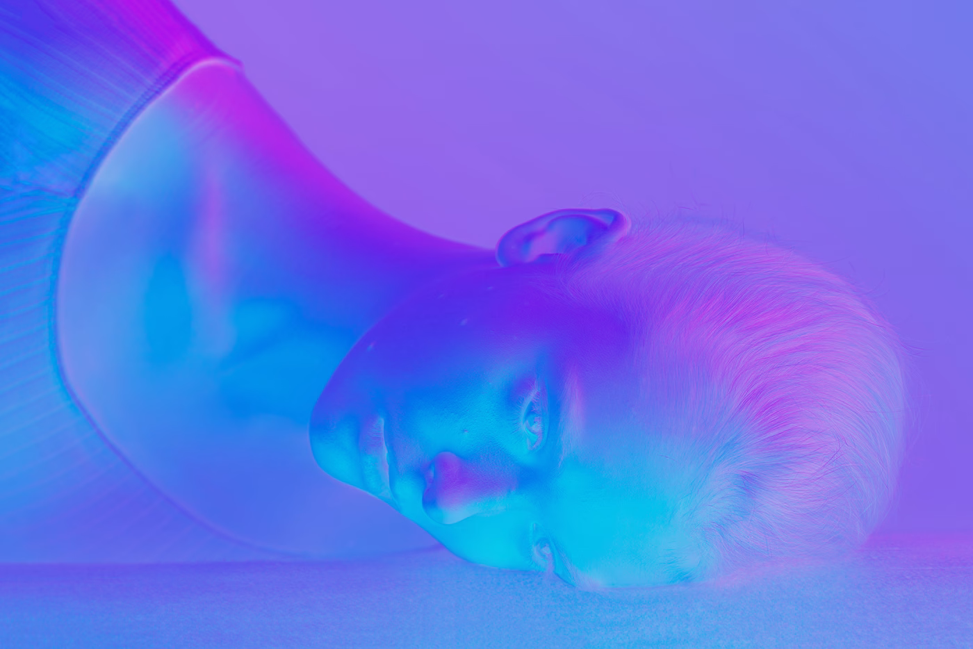 A close-up of a person lying on a surface, illuminated by vibrant blue and pink tones. Their expression is serene.