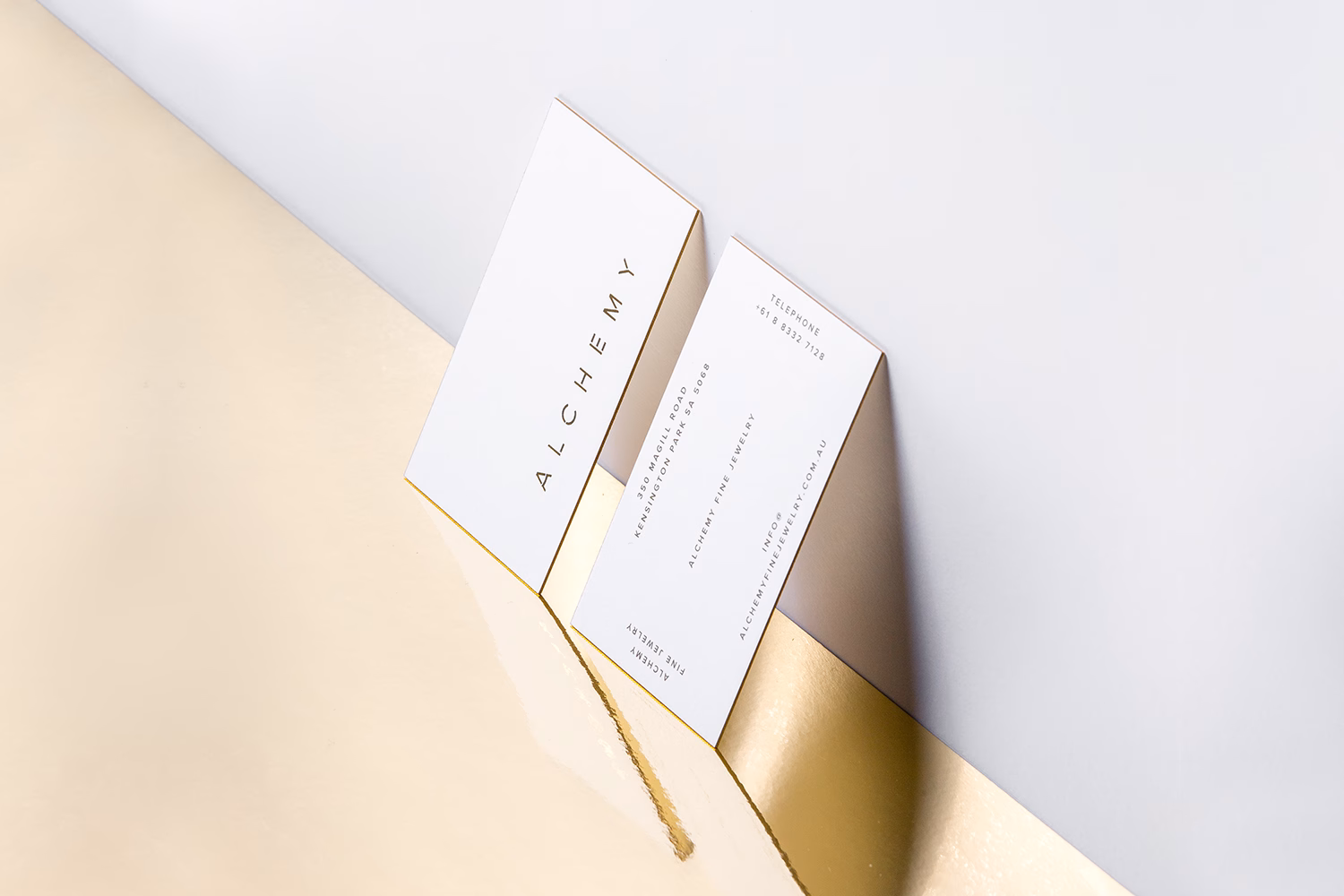 Two elegant, white business cards with gold edges rest against a reflective gold surface, showcasing minimalistic typography.