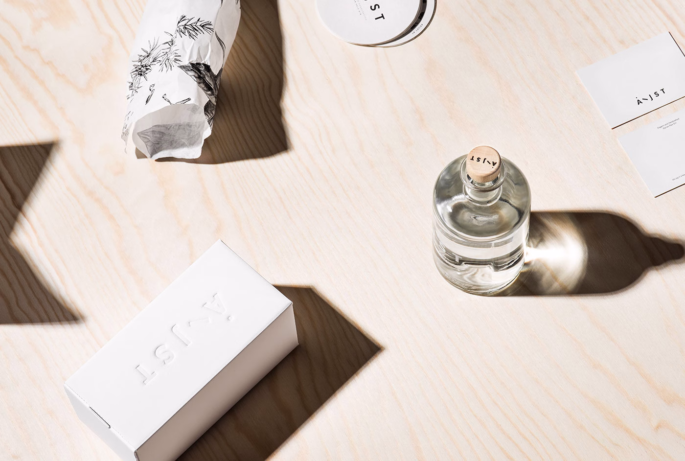 A glass bottle with a wooden cap sits on a light wood surface, accompanied by a white box and patterned wrapping paper.