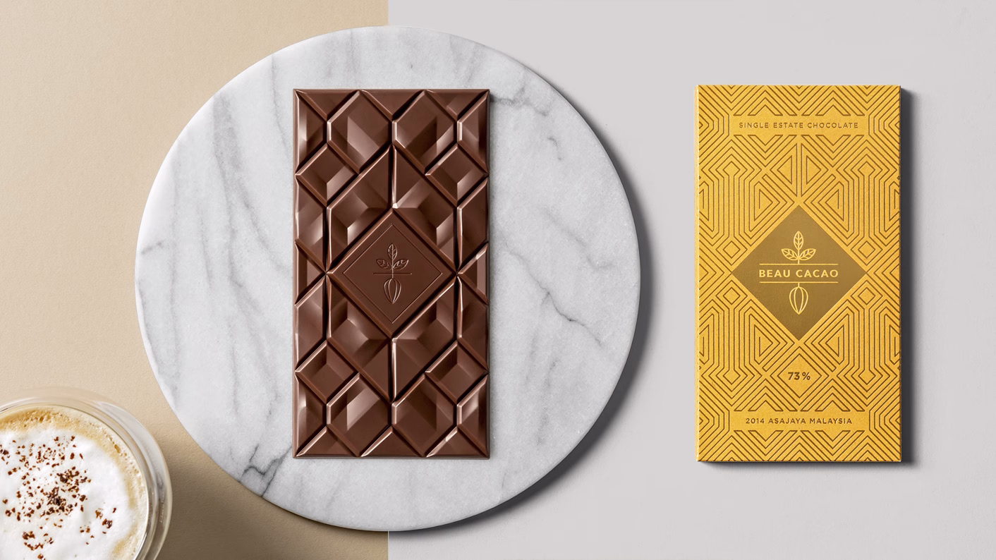 A marble plate holds a geometric chocolate bar beside a vibrant yellow wrapper, with a creamy beverage in the foreground.