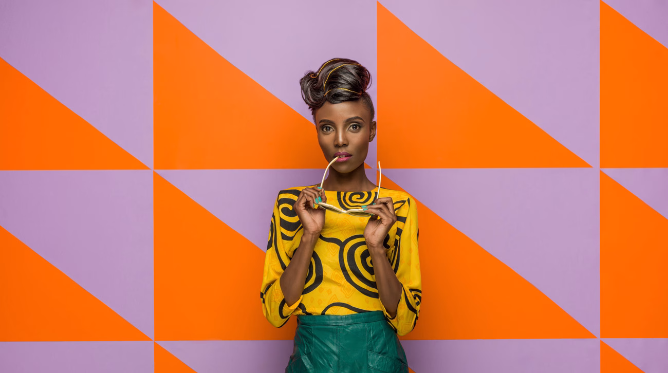 A person poses against a vibrant purple and orange geometric wall, wearing a colorful patterned top and holding sunglasses.