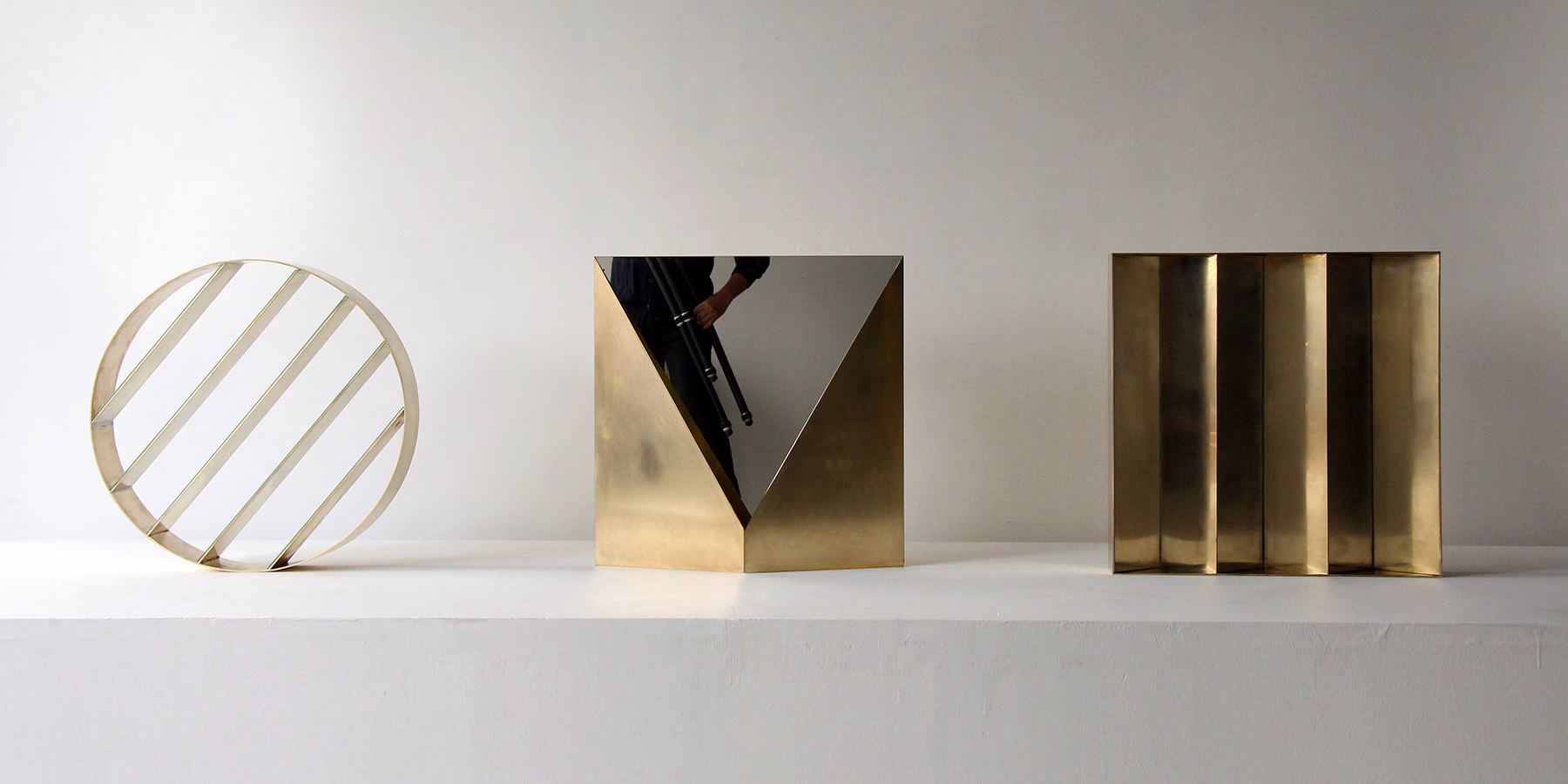 Three brass sculptures feature geometric forms: a circular frame with stripes, a triangular mirror, and a rectangular block.