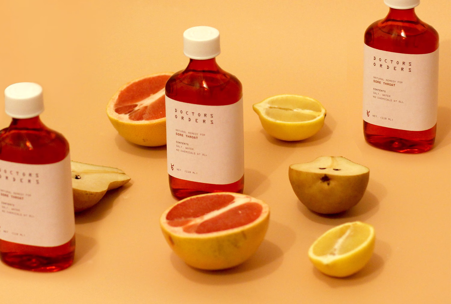 Three bottles of red liquid are surrounded by halved citrus fruits on a soft peach background.