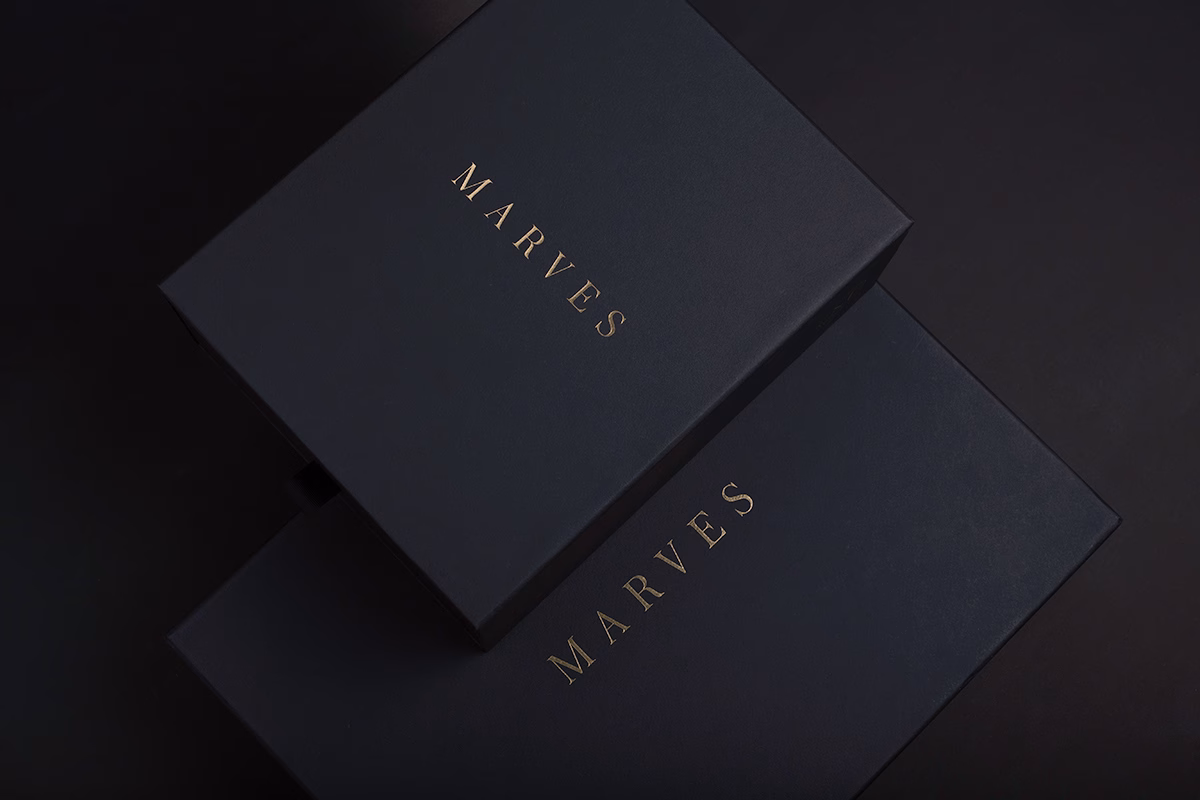 Two dark blue boxes with gold lettering spelling "MARVES," arranged at an angle on a black surface.