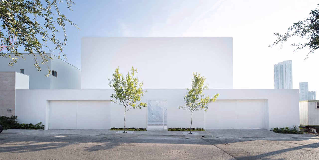 A minimalist white building facade is flanked by two small trees, creating a serene entrance against a clear sky.