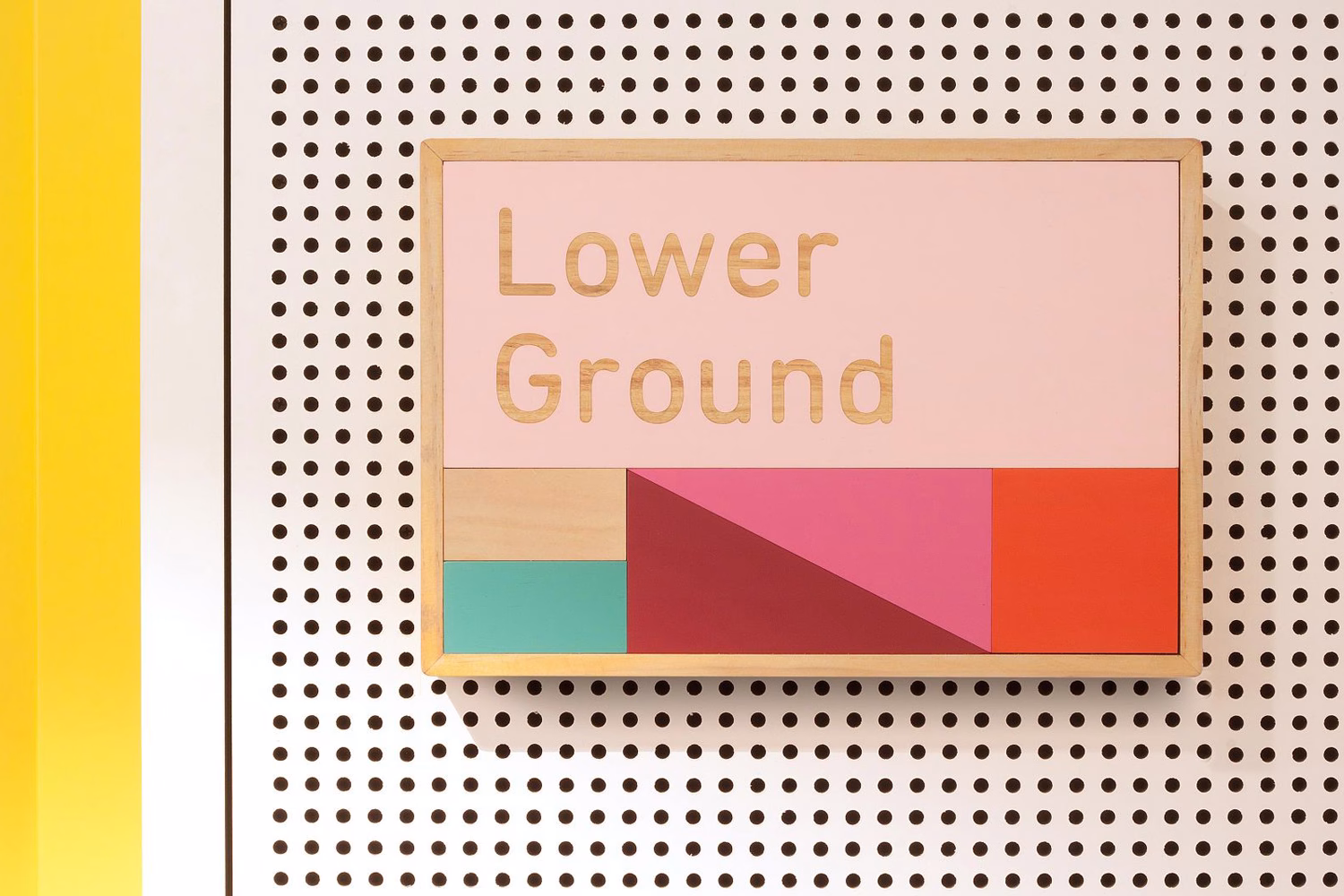 A wooden sign displaying "Lower Ground" in a light color, set against a perforated white wall and vibrant geometric shapes.