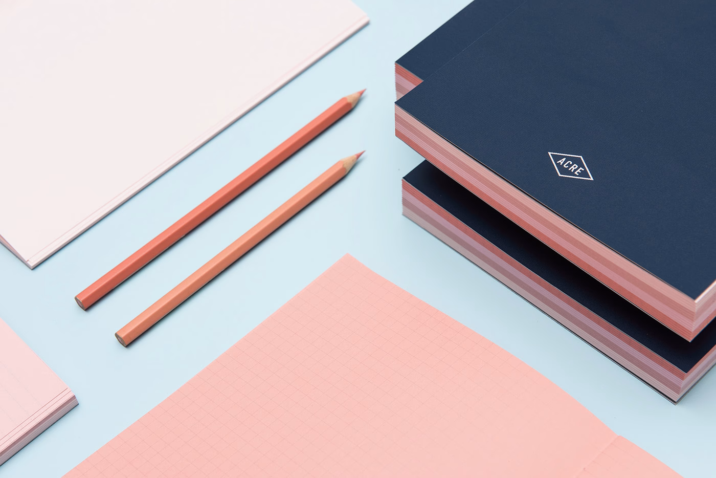 A pair of peach-colored pencils rests next to stacks of navy notebooks and pink sheets on a light blue surface.