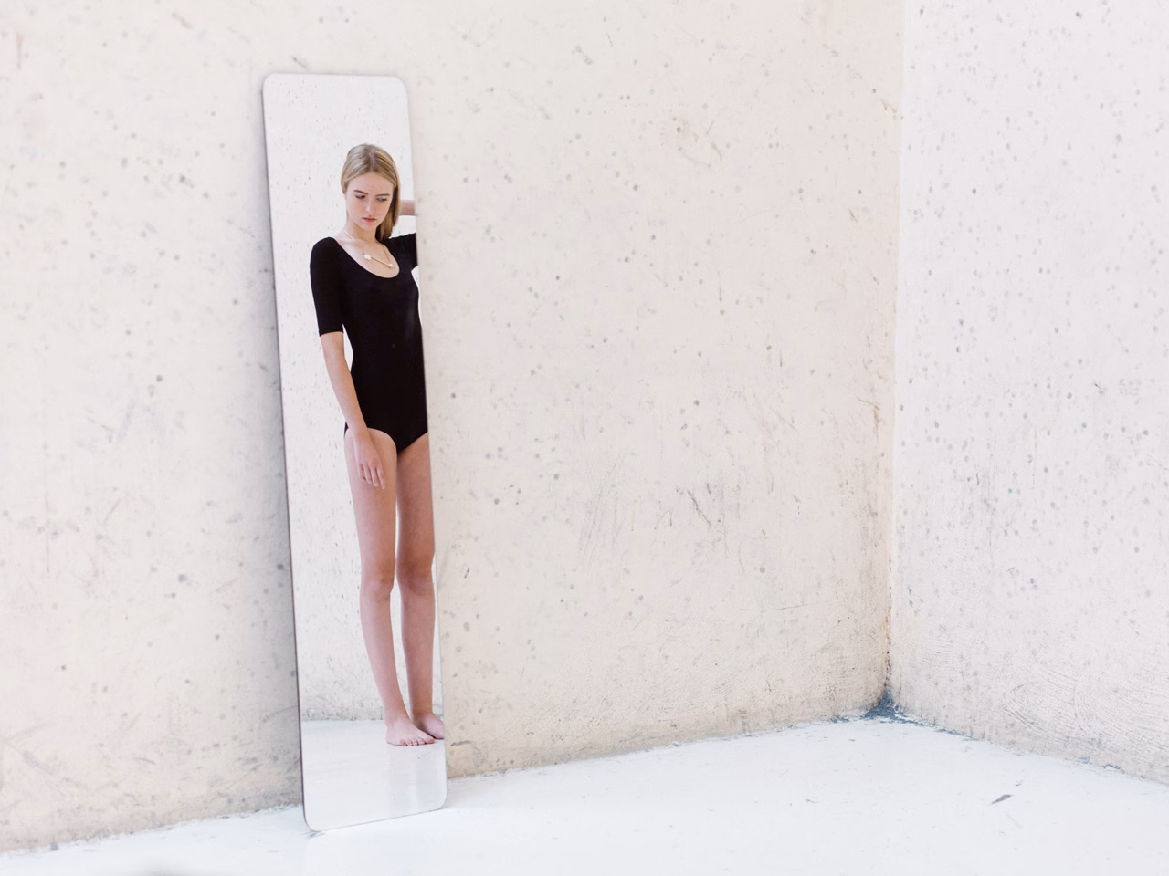 A young woman in a black bodysuit stands beside a tall, slim mirror against a neutral wall.