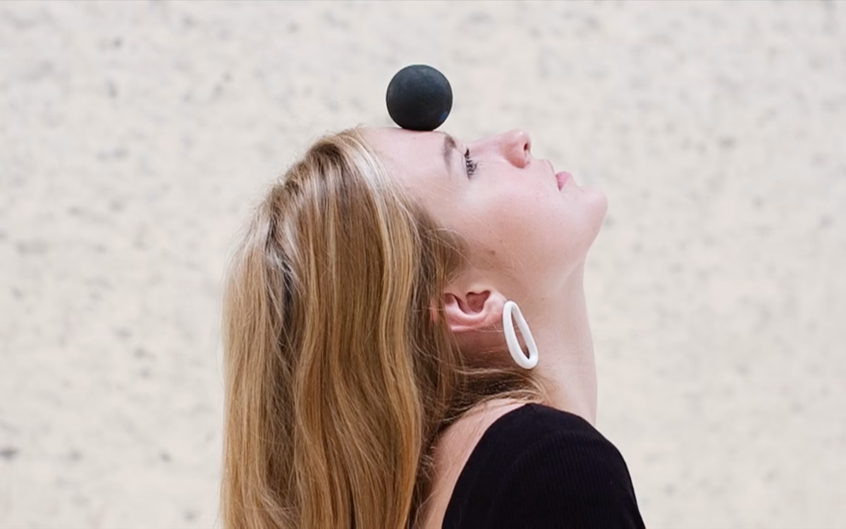 A woman with long hair tilts her head back, balancing a black sphere on her forehead against a pale background.
