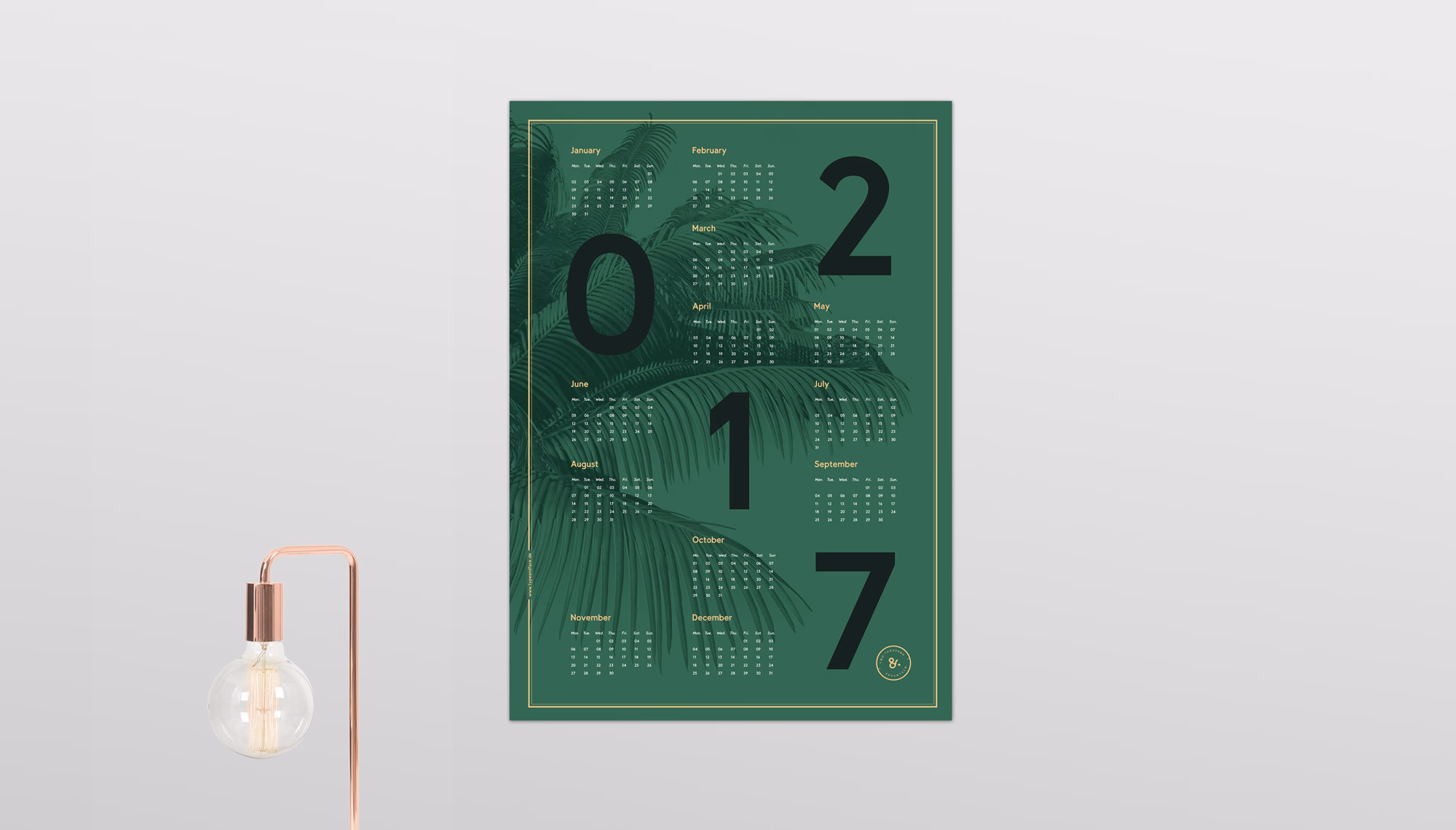 A green calendar features large black numbers for the year 2017, overlaid with palm leaves and a gold border. A copper lamp sits nearby.