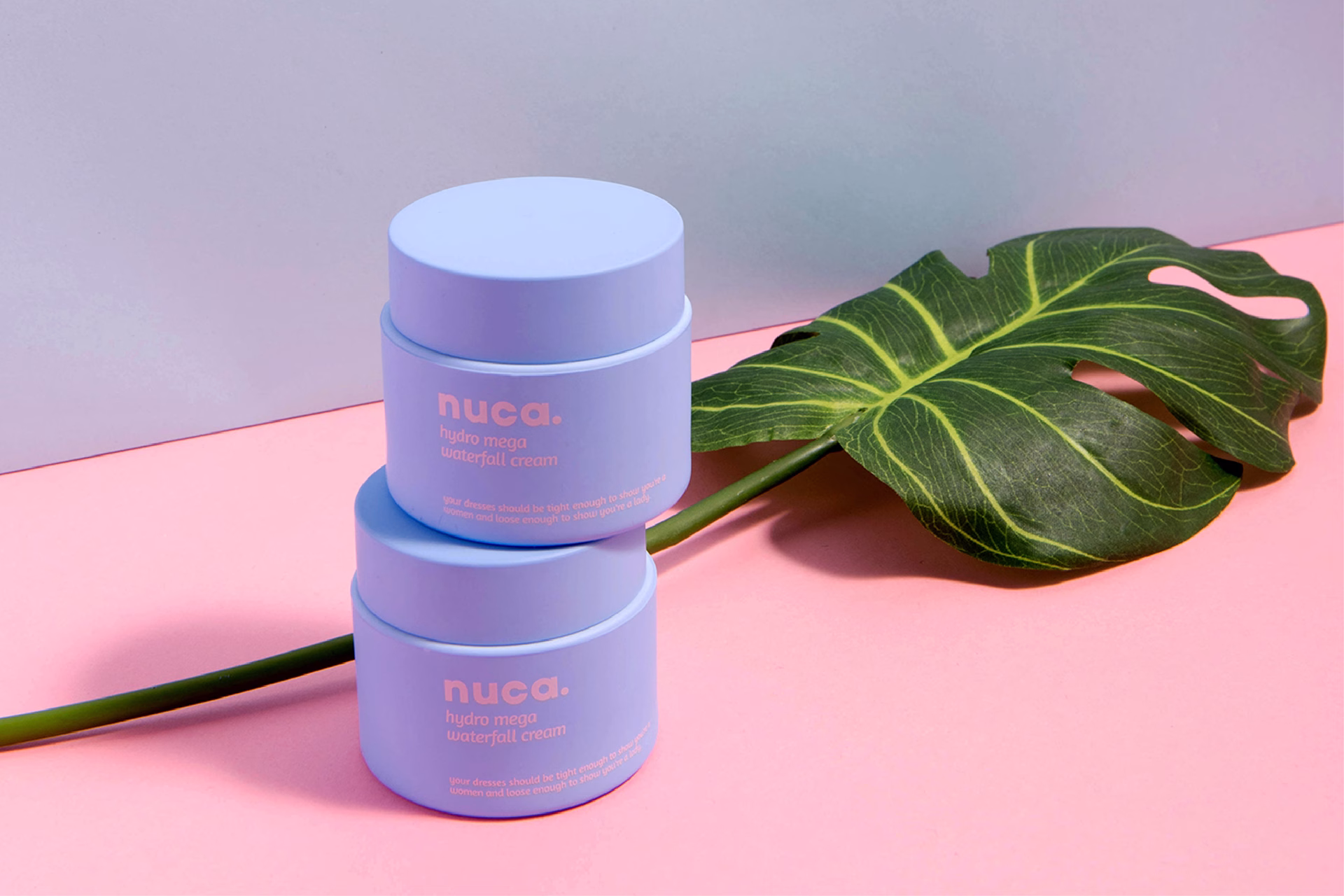 Two light blue cream jars labeled "nuca" sit atop a green leaf, against a pink background.