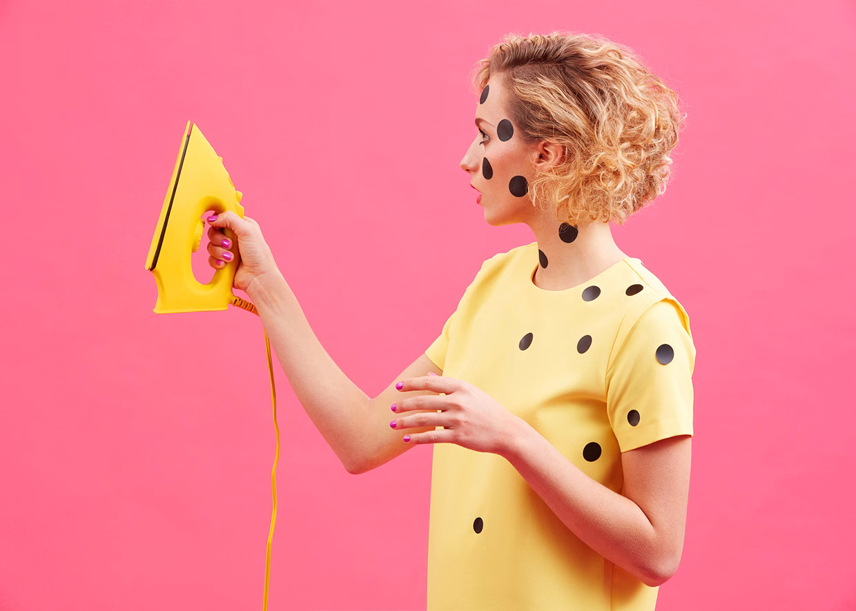 A person with curly hair holds a yellow iron, dressed in a yellow top with black polka dots against a pink background.