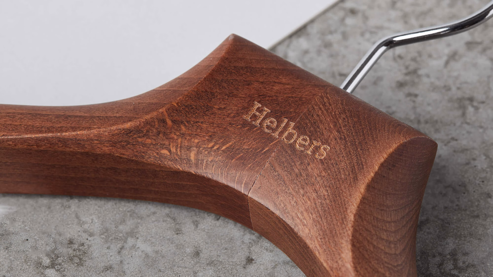 A polished wooden hanger with a subtle "Helbers" engraving rests on a textured gray surface.