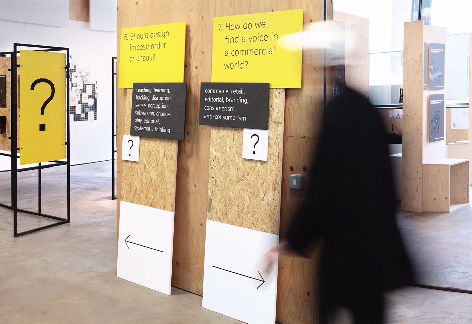 A blurred figure in a black coat passes by yellow and black panels with questions about design and commerce in a gallery space.
