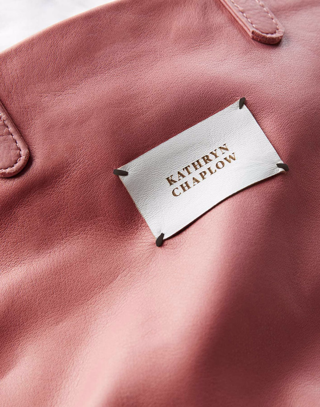 A close-up of a pink leather bag featuring a white label with the name "KATHRYN CHAPLOW" embossed in gold.
