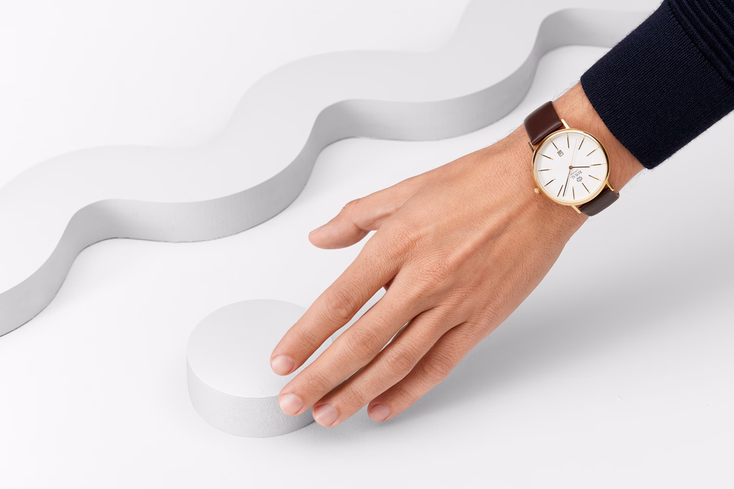 A hand reaches towards a white circular object on a smooth surface, wearing a gold watch with a brown leather band.