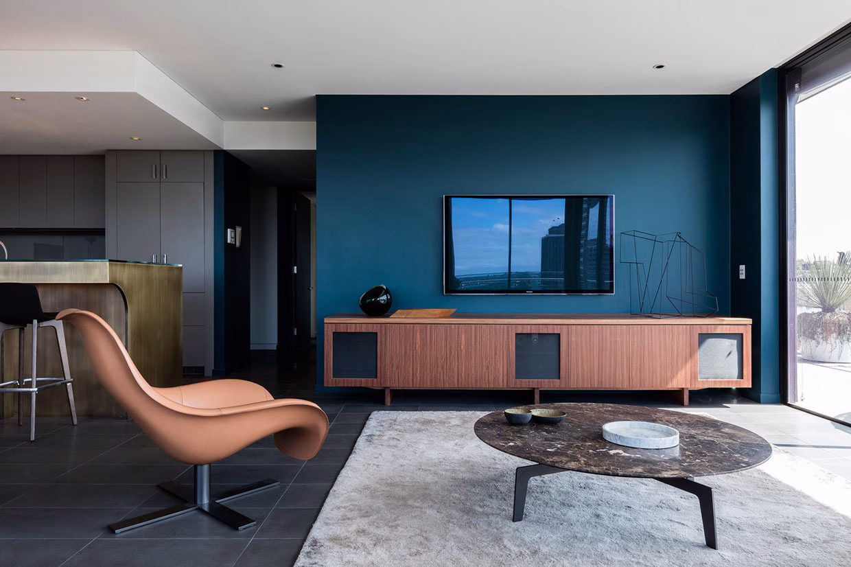 A modern living room features a curved brown chair, a wooden TV cabinet, and a marble coffee table on a light rug.