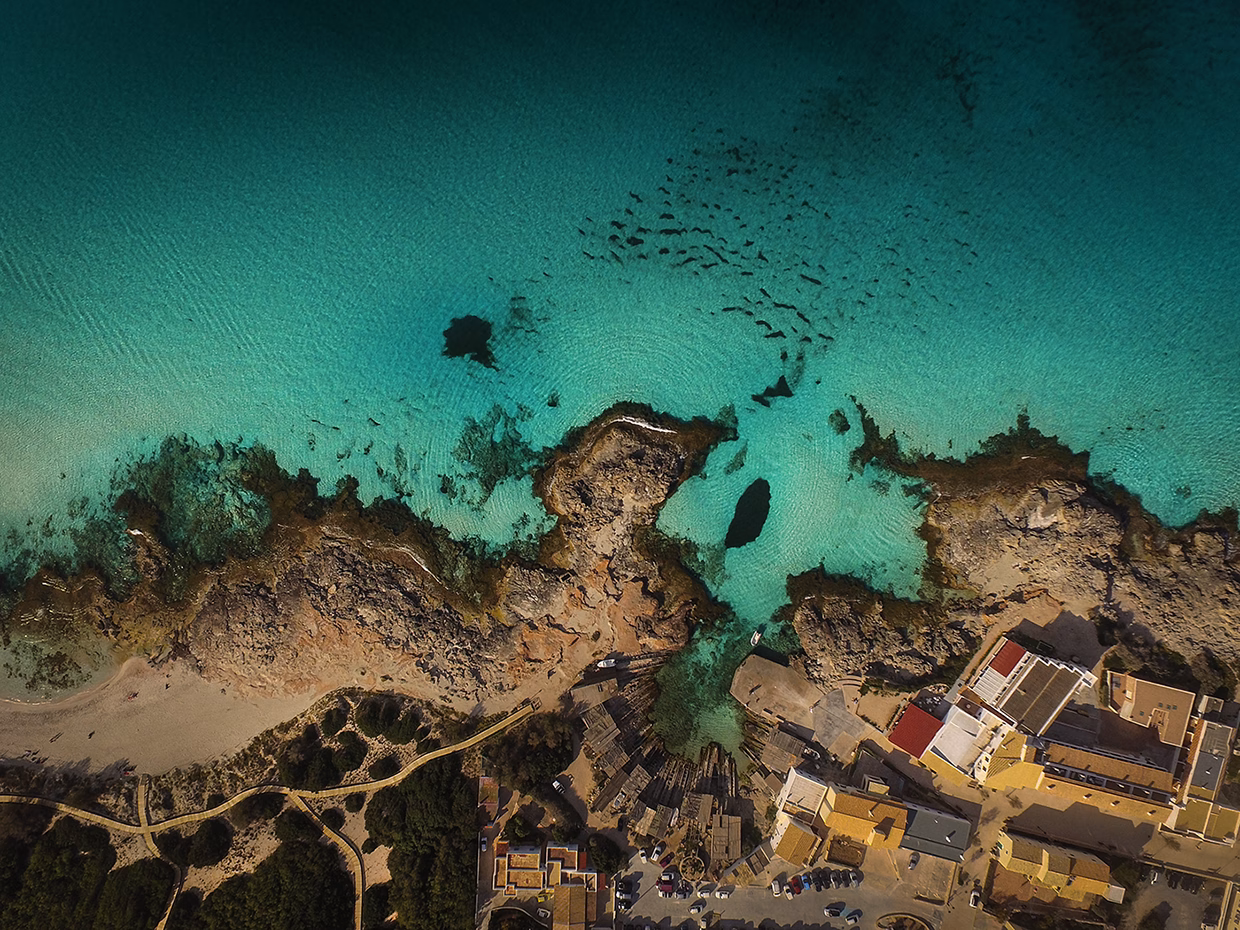 An aerial view of a turquoise bay, rocky shores, and a small coastal village nestled by the water's edge.