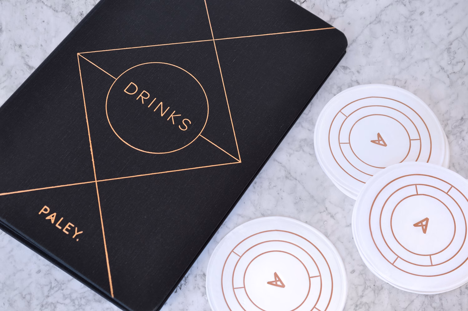 A black menu with gold typography reading "DRINKS" rests on a marble surface beside circular white coasters.