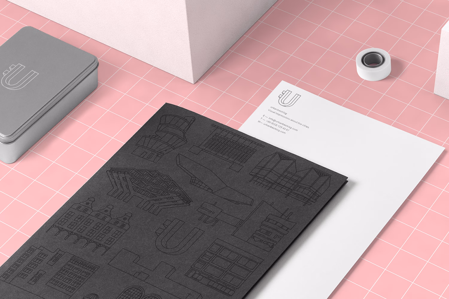 A black folder featuring architectural outlines rests on a pink grid surface, accompanied by a silver tin and a white tape.