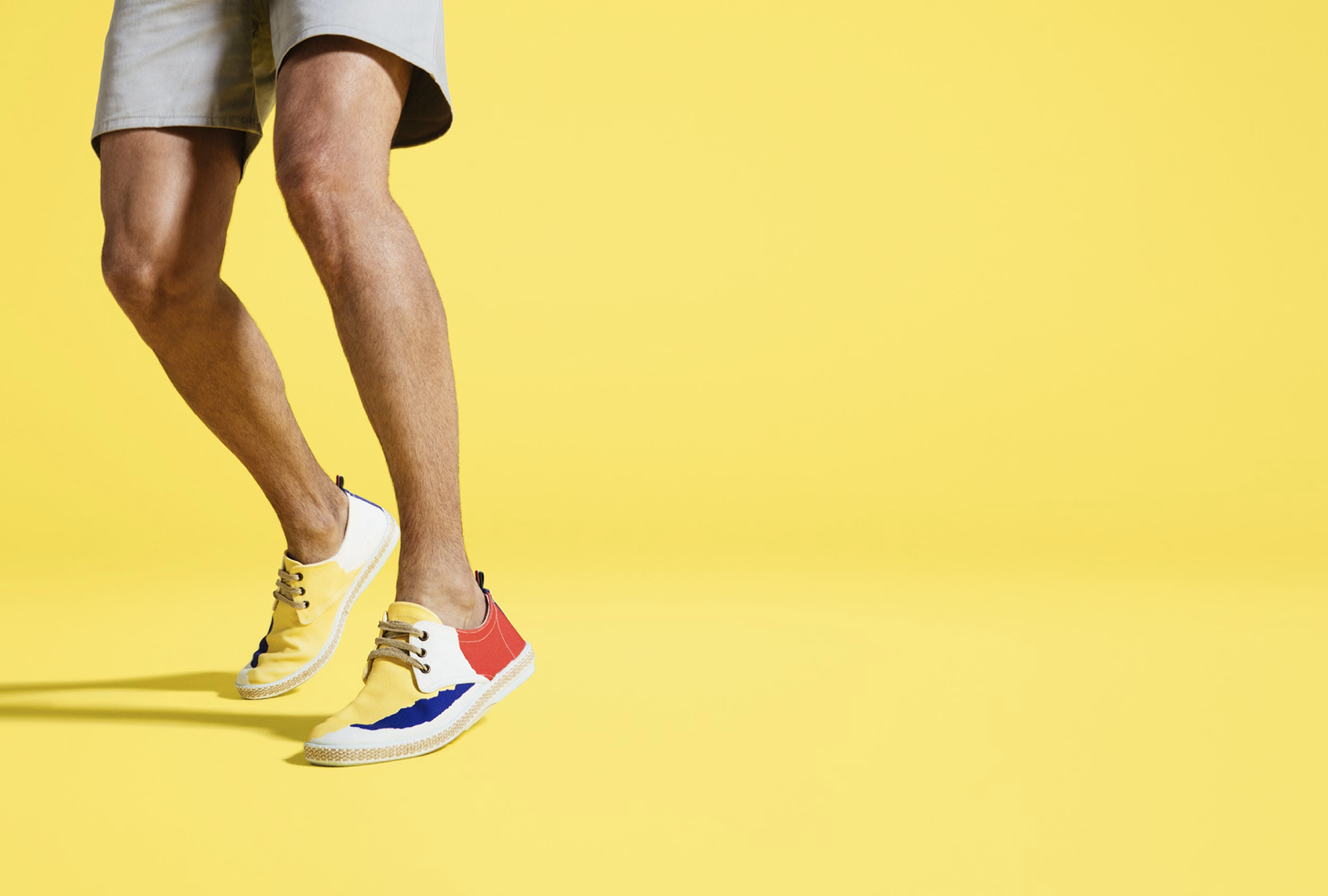 A person’s legs in light shorts are captured mid-step, showcasing colorful sneakers against a bright yellow background.