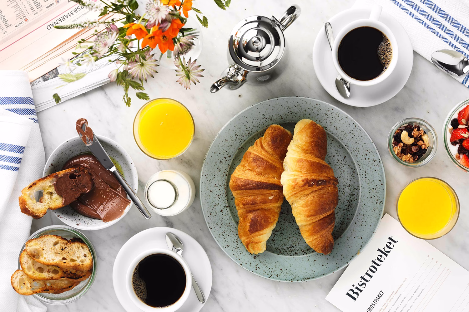 A spread of breakfast items including croissants, coffee, and orange juice on a marble table, accented by flowers.