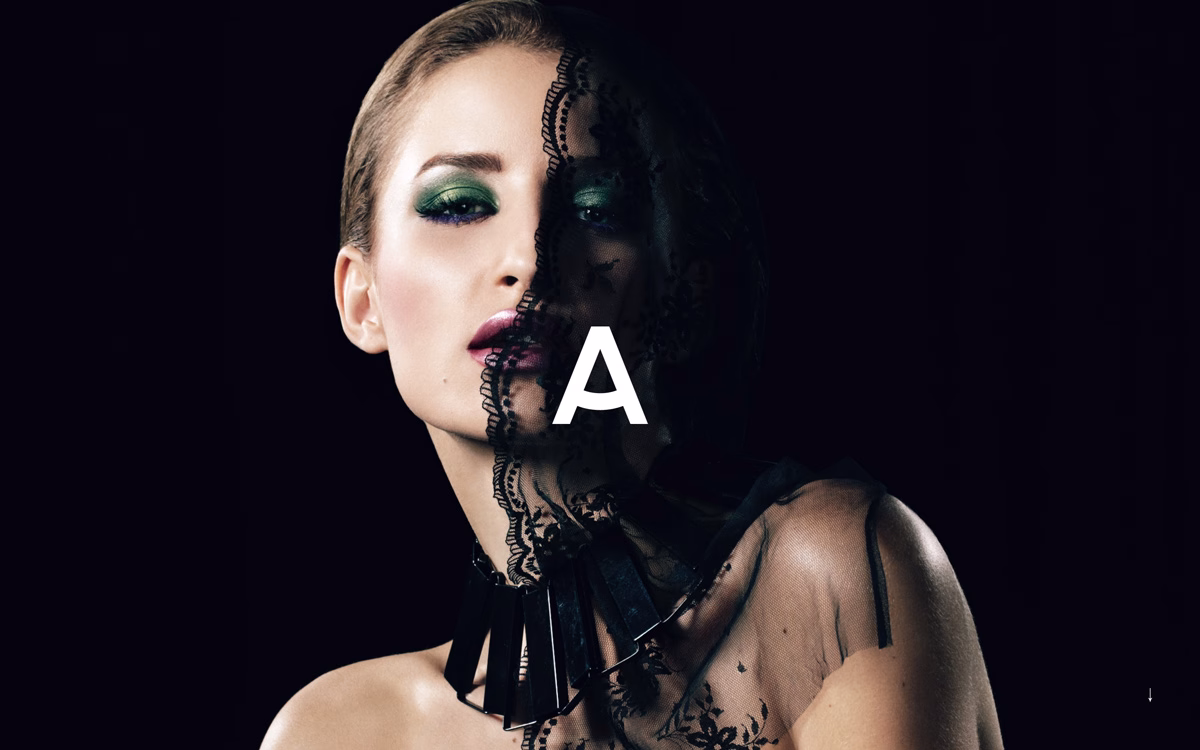 A woman with bold green eyeshadow and deep burgundy lips poses in a dramatic black lace outfit against a dark backdrop.
