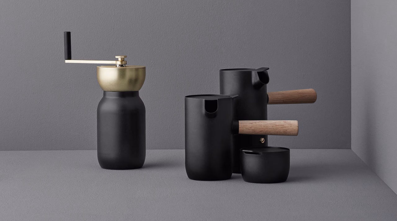 A black ceramic coffee grinder and various vessels with wooden handles arranged on a minimal gray surface.