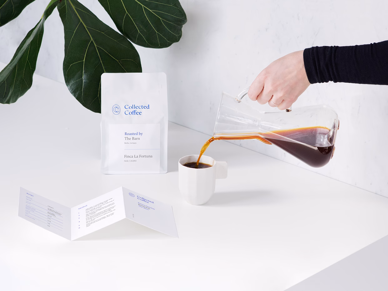 A hand pours coffee from a glass carafe into a white cup, alongside a bag of Collected Coffee on a light surface.