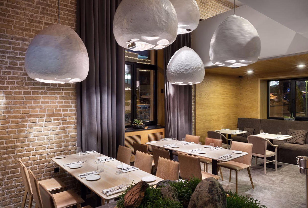 A modern restaurant interior featuring wooden tables, soft lighting from sculptural pendant lamps, and grey curtains.