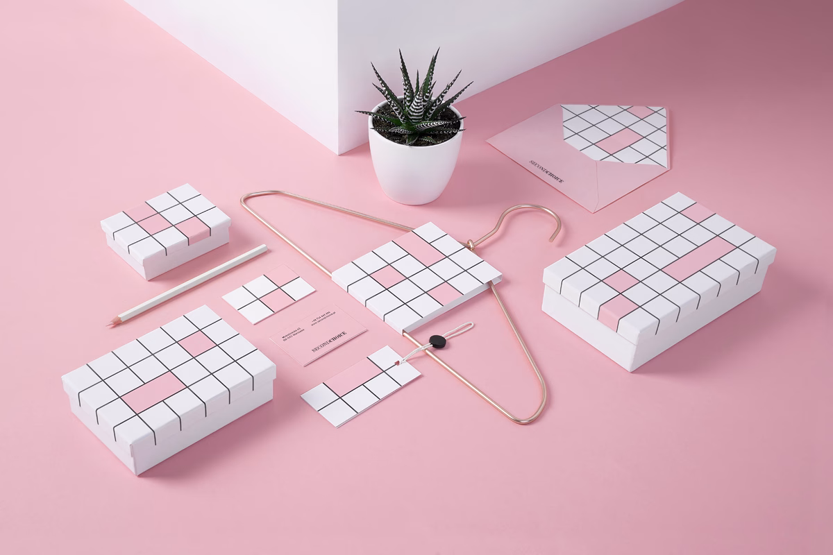 A collection of white packaging with pink accents, a pencil, and a small potted plant arranged on a pink surface.