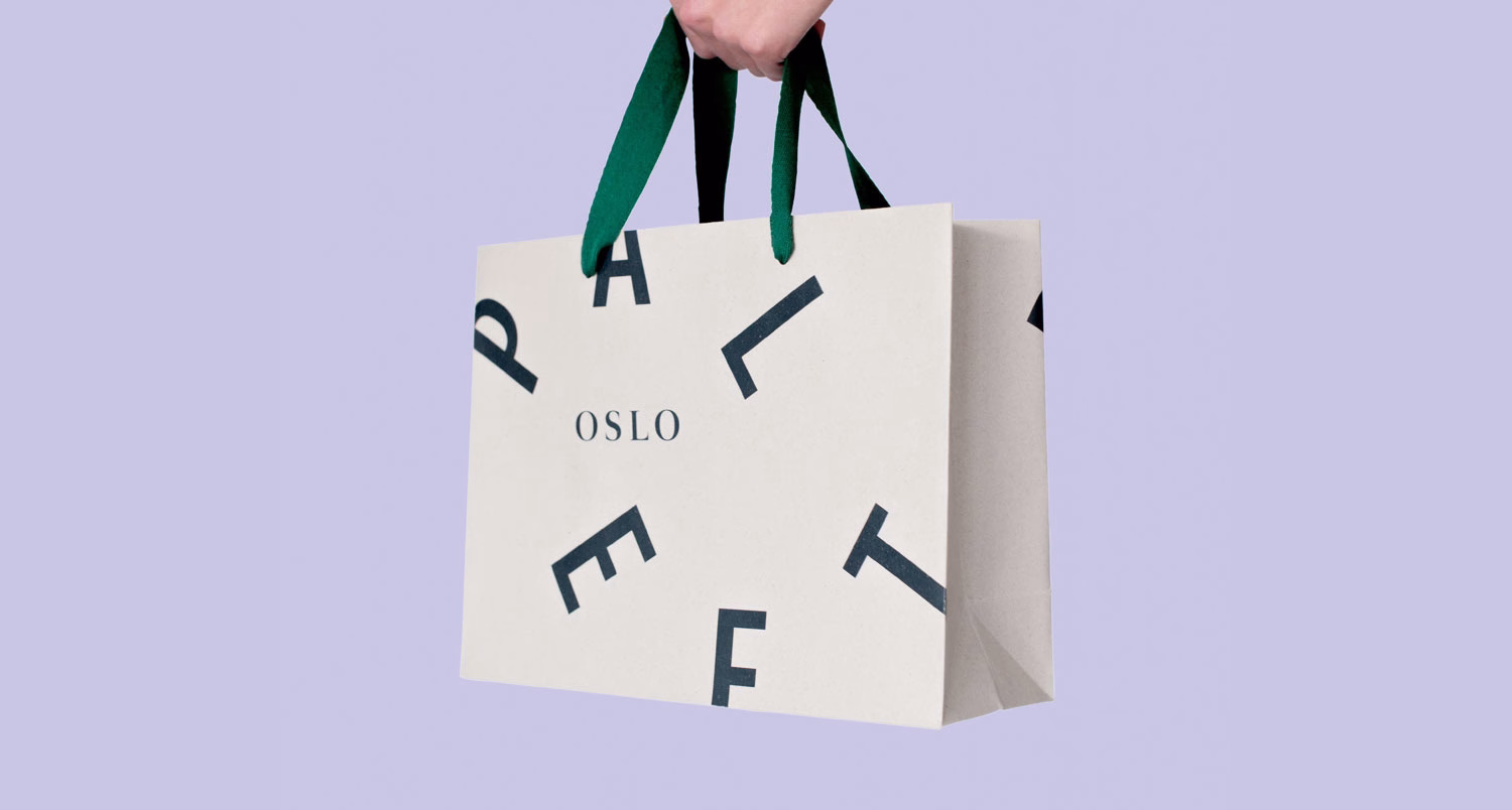 A hand holds a cream shopping bag with dark green handles, featuring scattered black letters and the word "OSLO."