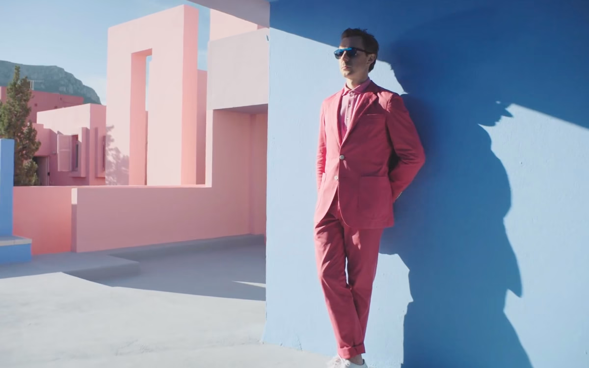 A man in a pink suit leans against a blue wall, surrounded by pastel-colored architectural structures.
