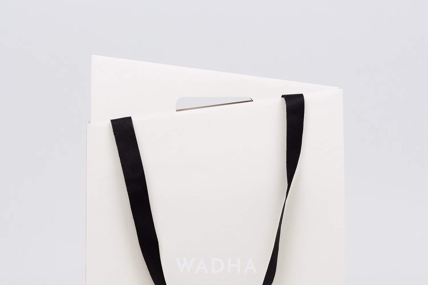 A white paper bag with black handles displays the word "WADHA" in simple, modern typography.